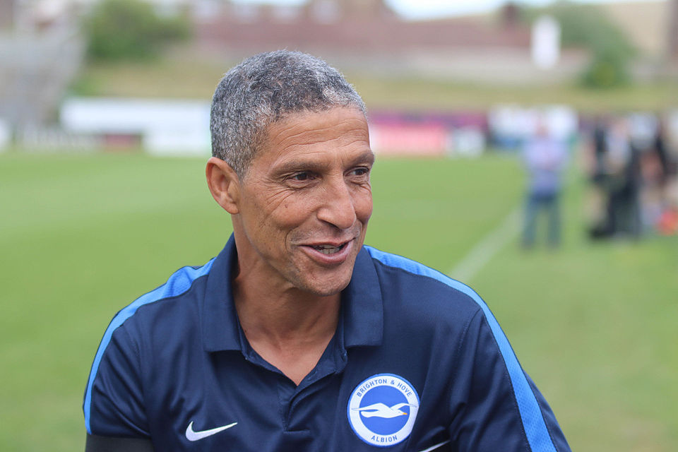 A smiling man with black & grey hair and a blue shirt. The shirt has a Brighton & Hove Albion logo. In the background is a football pitch.